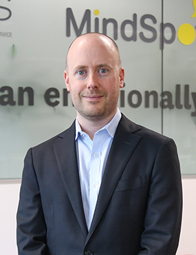 Associate Professor Blake Dear, a smiling man in a suit, standing in front of a MindSpot sign.