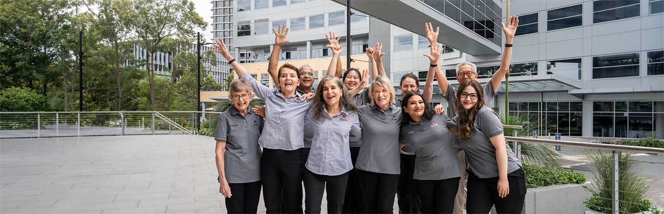A group of ten volunteers in matching grey polo shirts, smiling and waving their hands in the air.