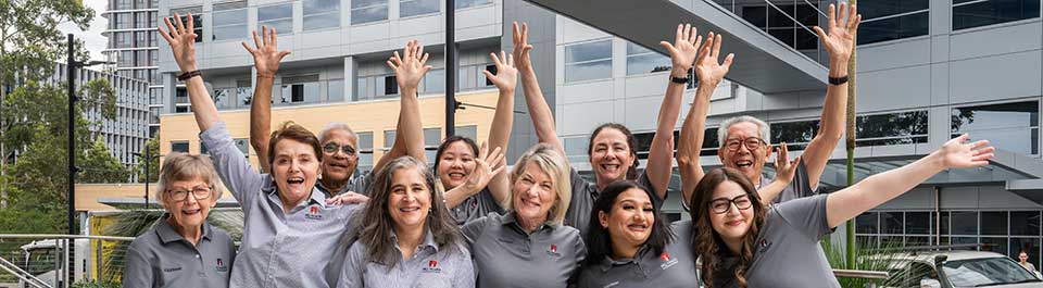 A group of ten volunteers in matching grey polo shirts, smiling and waving their hands in the air.