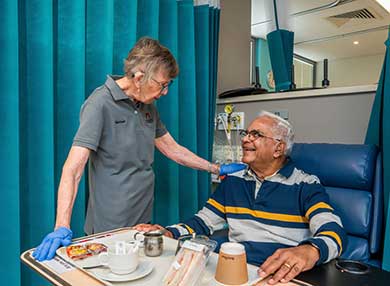 A volunteer in a grey polo shirt chatting with a smiling patient over their lunch tray.